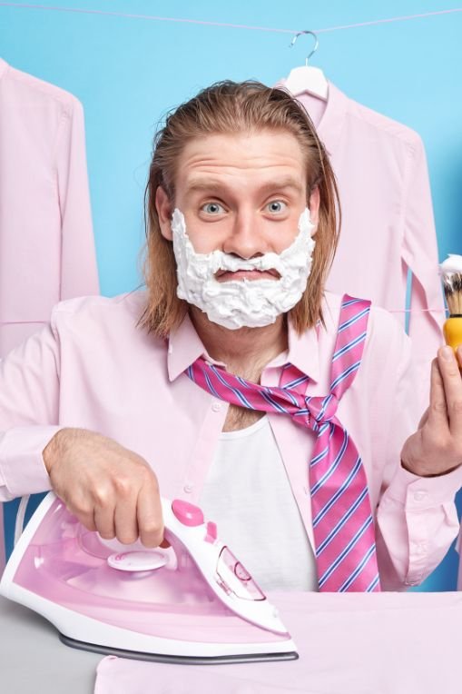 Man with shaving cream on his face ironing a shirt while wearing a pink tie – concept for meme marketing humour.